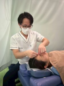 高幡不動院の施術室で、花粉症に悩む女性の眉間周辺に鍼を打つスタッフ。マスクとメガネを着用したスタッフが、丁寧に鍼灸施術を行っている様子。 